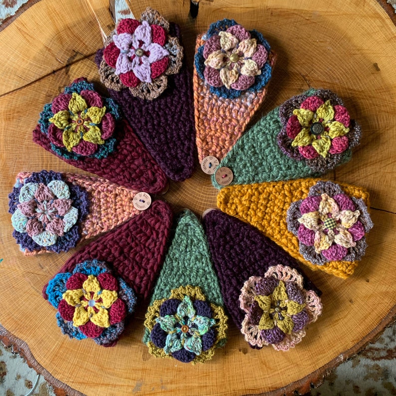 May include: A collection of eight triangular crochet headbands arranged in a circle. Each headband features a unique color combination of yarn and a large, multi-layered crocheted flower embellishment attached to the center with a small button.