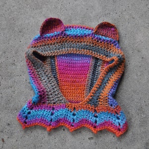 May include: A colorful crocheted hooded scarf with ear flaps. The scarf is made with a variety of colors, including orange, pink, blue, and gray.