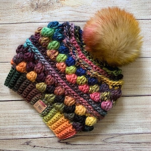 May include: A colourful crocheted beanie with a faux fur pom pom. The beanie is made with a variety of colours, including red, orange, yellow, green, blue, purple, and brown. The beanie has a small tag that says "Of Mars".