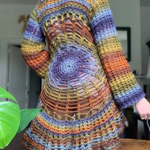 May include: A long-sleeved crochet tunic featuring a vibrant, multi-coloured gradient yarn in shades of orange, yellow, blue, and purple. The garment showcases an intricate, open-work spiderweb pattern across the back, transitioning into a flared hem.