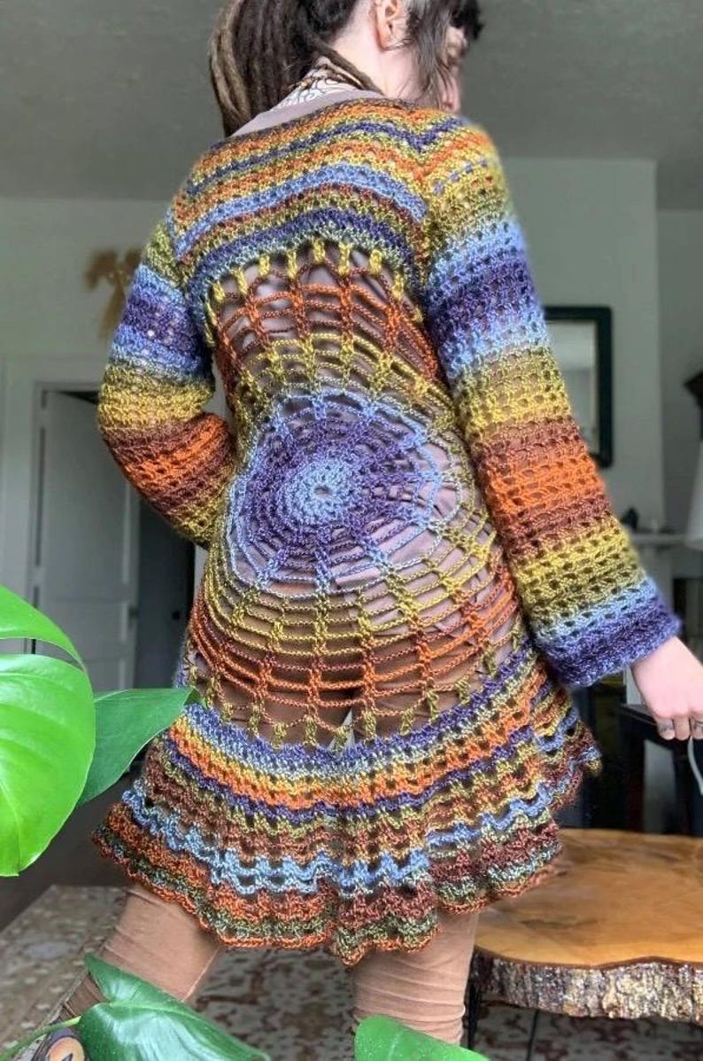 May include: A long-sleeved crochet tunic featuring a vibrant, multi-coloured gradient yarn in shades of orange, yellow, blue, and purple. The garment showcases an intricate, open-work spiderweb pattern across the back, transitioning into a flared hem.