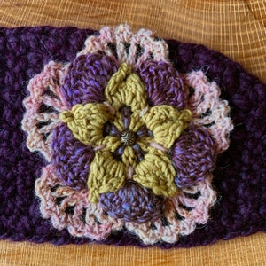 May include: A dark purple crochet headband featuring a multi-layered floral applique. The flower has mustard yellow inner petals, purple middle petals, and a light pink lacy outer border, all centered with a small, round, textured metallic bead.