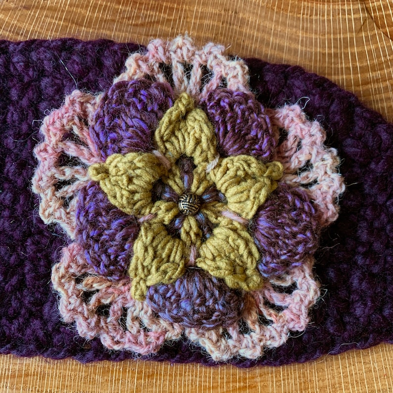 May include: A dark purple crochet headband featuring a multi-layered floral applique. The flower has mustard yellow inner petals, purple middle petals, and a light pink lacy outer border, all centered with a small, round, textured metallic bead.