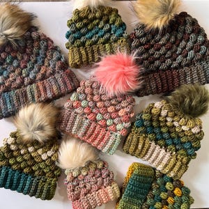 Może przedstawiać: Kolekcja dziesięciu czapek na szydełku w różnych kolorach i wzorach. Czapki są wykonane z fakturowanym ściegiem, który tworzy nierówną powierzchnię. Każda czapka ma puszysty pompon na górze.