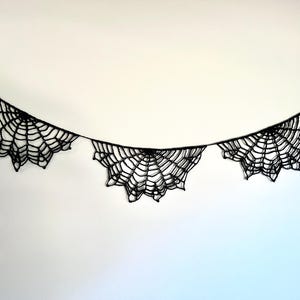 Op de afbeelding: Een zwarte spinnenweb slinger, ideaal voor Halloween decoraties. De slinger heeft drie web ontwerpen verbonden door een dun zwart touwtje. De webben zijn ingewikkeld ontworpen met een gedetailleerd patroon, wat een spookachtige uitstraling cre&euml;ert.