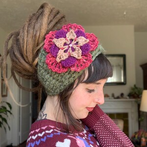 May include: A wide, olive green crocheted headband featuring a large, multi-layered floral applique. The flower is constructed from concentric layers of dark purple, bright pink, and light tan yarn, with a small central bead detail.