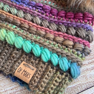 Puede incluir: Un gorro de crochet marrón con un patrón de rayas multicolores y un pompón en la parte superior. El gorro tiene una etiqueta de cuero marrón con el texto "Of Mars" en ella.