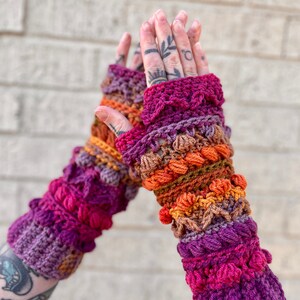 May include: A pair of colorful crocheted fingerless gloves with a textured design. The gloves are made with a variety of shades of purple, orange, and brown yarn.