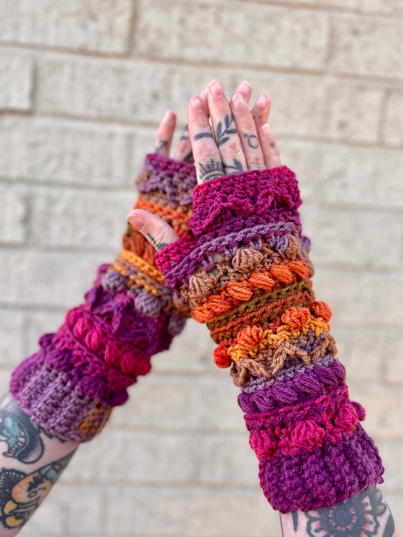 May include: A pair of colorful crocheted fingerless gloves with a textured design. The gloves are made with a variety of shades of purple, orange, and brown yarn.