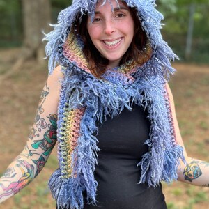 May include: A blue, yellow, and pink crocheted hooded scarf with fringe. The scarf is worn by a person with tattoos on their arms.