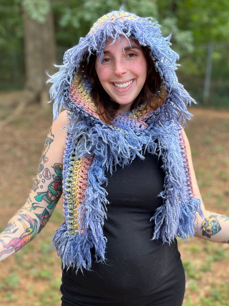 May include: A blue, yellow, and pink crocheted hooded scarf with fringe. The scarf is worn by a person with tattoos on their arms.