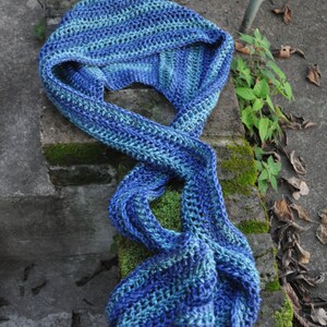May include: A blue and green crocheted scarf with a striped pattern. The scarf is folded and draped over a stone surface with moss growing on it.