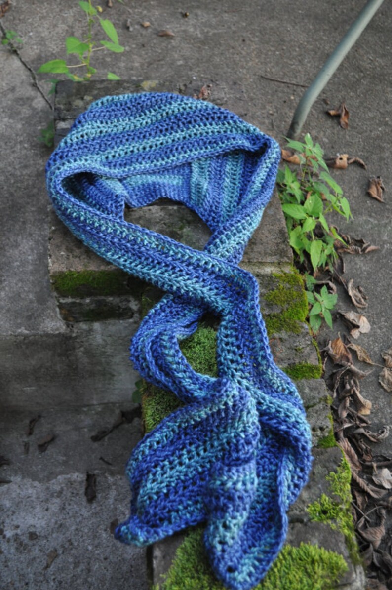 May include: A blue and green crocheted scarf with a striped pattern. The scarf is folded and draped over a stone surface with moss growing on it.
