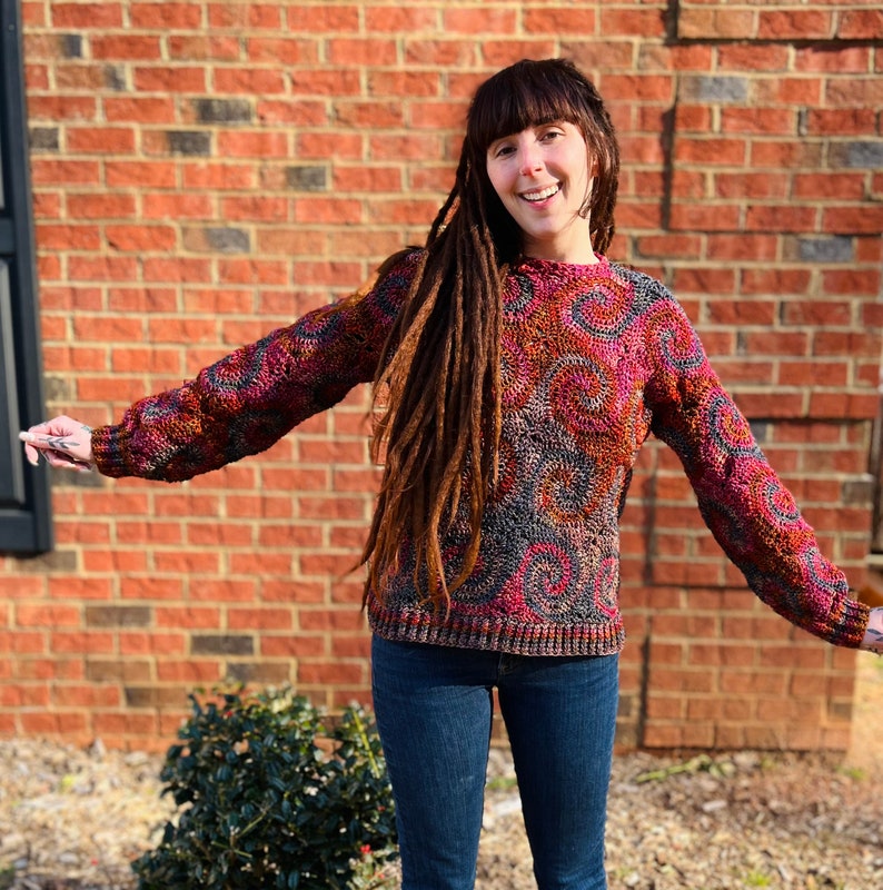 May include: A woman wearing a colourful crocheted sweater with a swirling pattern. She has long brown dreadlocks and is wearing blue jeans.
