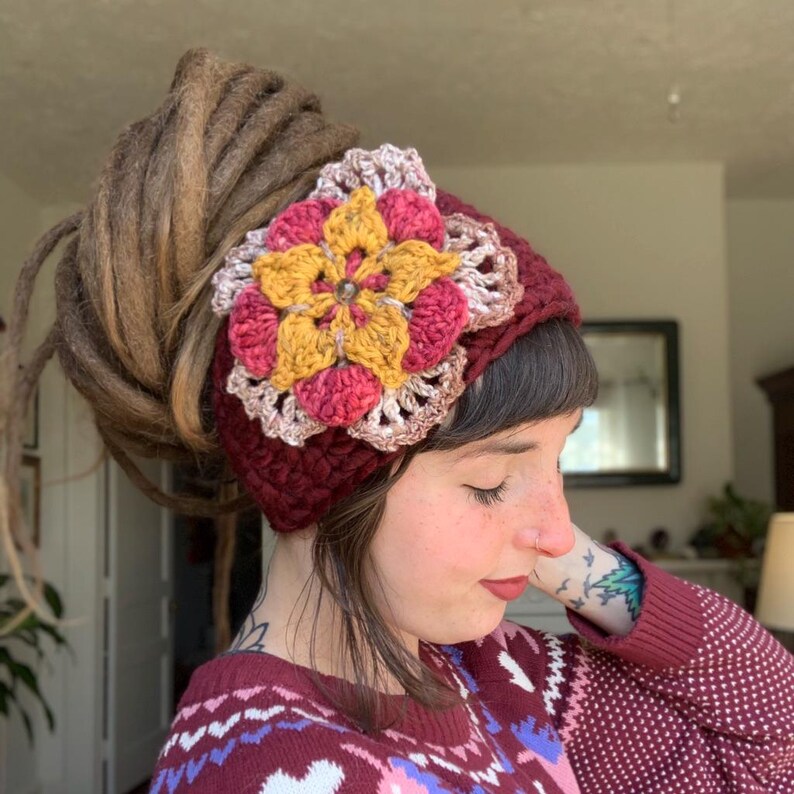 May include: A dark burgundy crochet headband featuring a large, multi-layered floral applique. The flower is crafted with yellow, pink, and light brown yarn, centered with a small, clear, faceted bead. The headband has a thick, textured knit.
