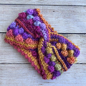 May include: A crocheted headband with a twisted design. The headband is made with a variety of colors, including purple, pink, orange, yellow, and green. The headband has a textured, bumpy surface.