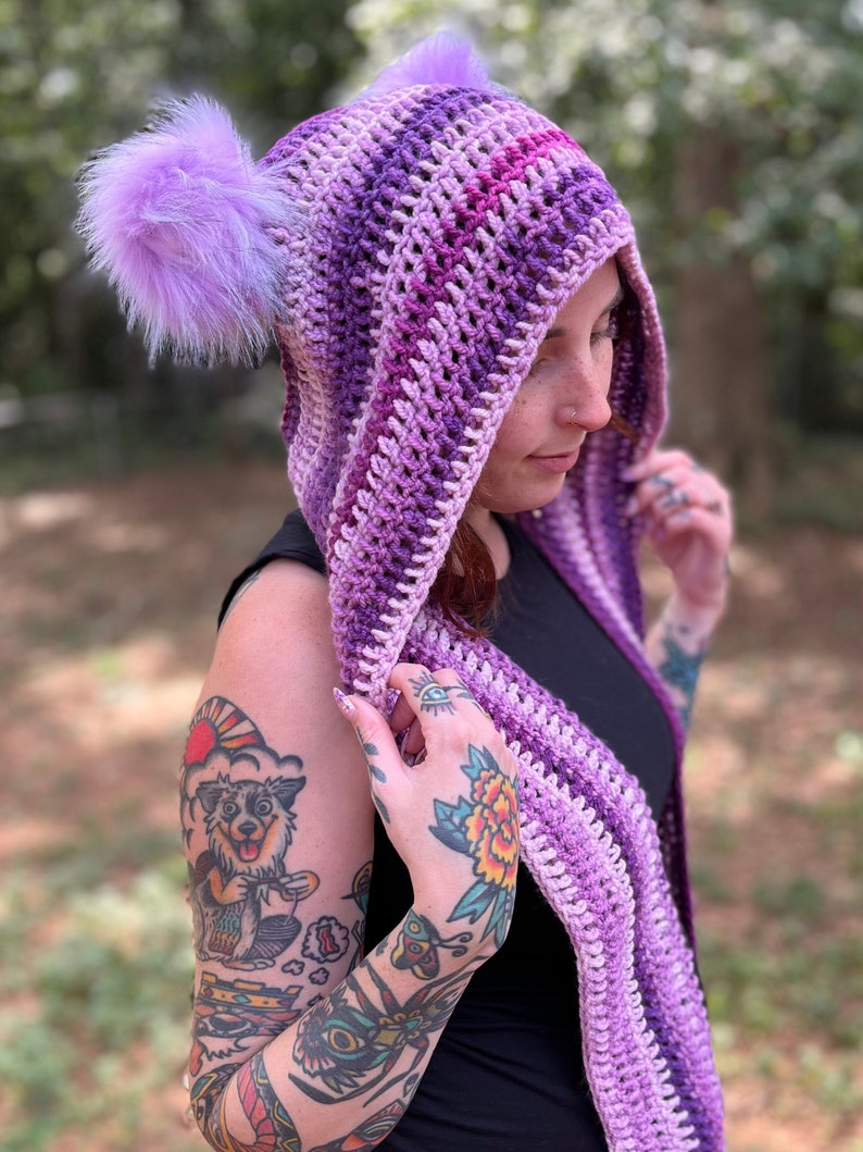 May include: A purple and pink striped crocheted hooded scarf with two fluffy pom-poms on the hood.
