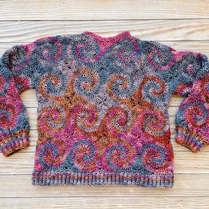 May include: A crocheted sweater with a colourful swirl pattern in shades of pink, orange, brown, and grey. The sweater has long sleeves and a crew neckline.