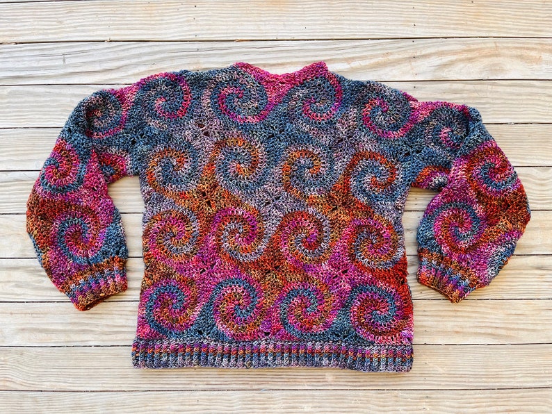May include: A crocheted sweater with a colourful swirl pattern in shades of pink, orange, brown, and grey. The sweater has long sleeves and a crew neckline.