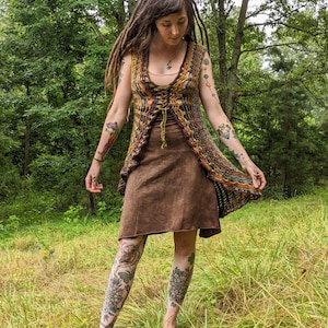 May include: A sleeveless, open-front crochet vest featuring a multi-colored pattern of orange, yellow, blue, and brown yarn with a lace-up front closure, worn over a solid brown knee-length dress with a simple scoop neckline and a straight hem.
