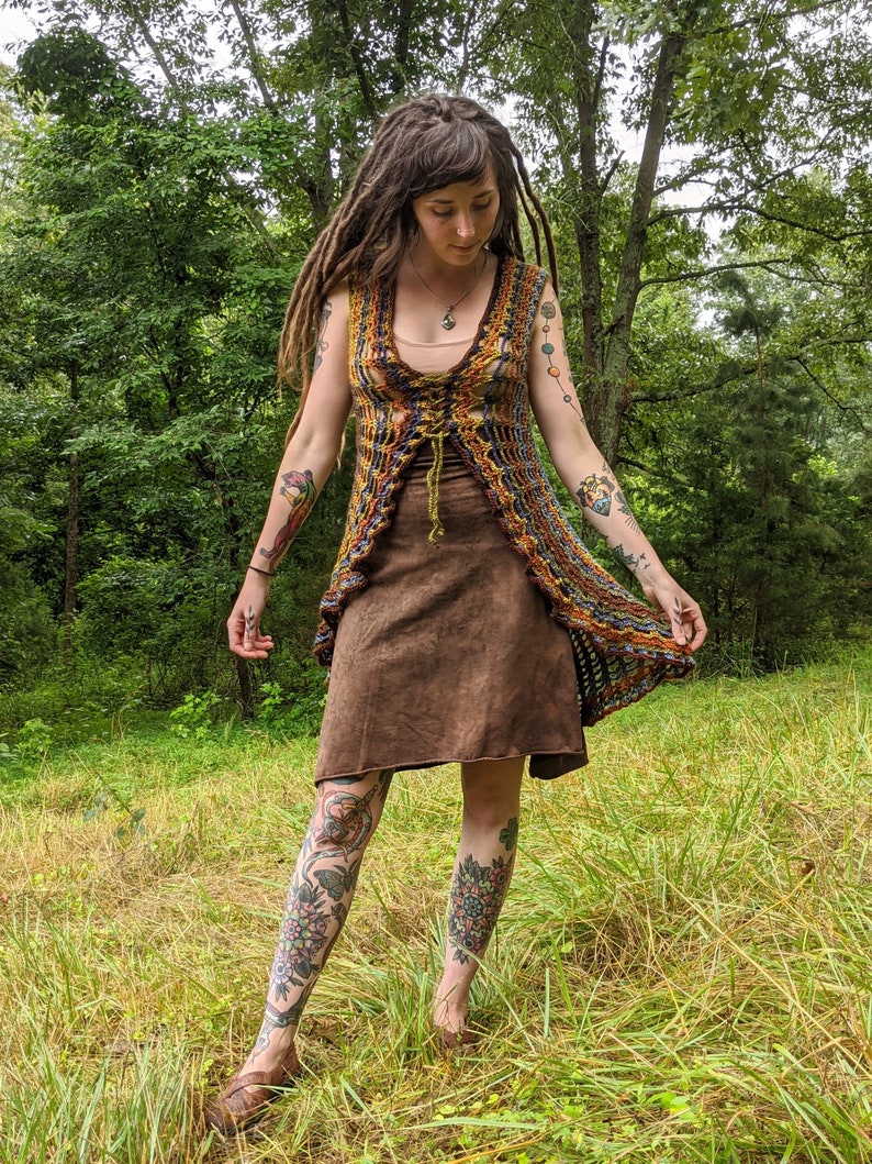 May include: A sleeveless, open-front crochet vest featuring a multi-colored pattern of orange, yellow, blue, and brown yarn with a lace-up front closure, worn over a solid brown knee-length dress with a simple scoop neckline and a straight hem.