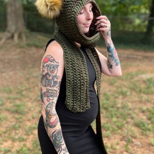 May include: A green crocheted hooded scarf with a brown pom-pom. The scarf is worn by a person with tattoos on their arms.