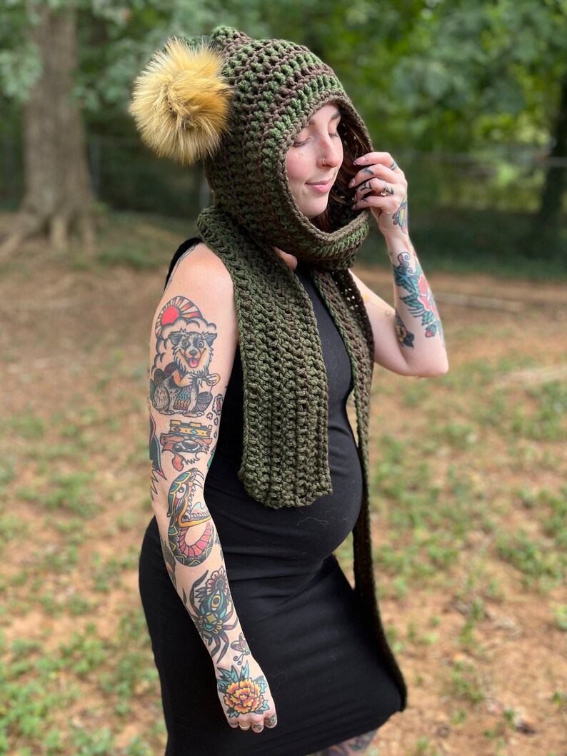 May include: A green crocheted hooded scarf with a brown pom-pom. The scarf is worn by a person with tattoos on their arms.
