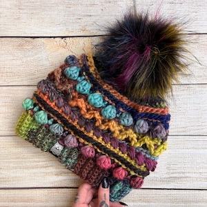 Puede incluir: Un gorro de punto de crochet colorido con un gran pompón esponjoso. El gorro está hecho con una variedad de texturas y colores, incluyendo verde, azul, morado, naranja y marrón. El pompón es negro con vetas moradas y amarillas.
