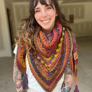 Può includere: Uno scialle all'uncinetto fatto a mano dai colori vivaci, arancione, giallo, rosa e viola. Lo scialle triangolare presenta un motivo strutturato e nappe decorative. I colori del filato si fondono per creare un accessorio sorprendente. Un articolo unico, fatto a mano.