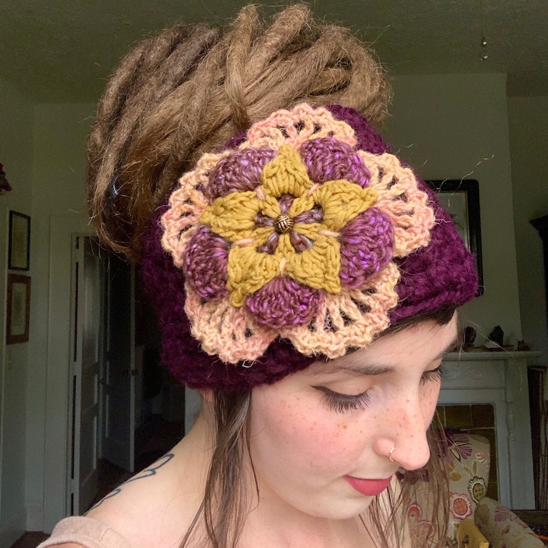 May include: A deep purple crochet headband featuring a large, multi-layered floral applique. The flower center is mustard yellow with a small metallic bead, surrounded by dark purple petals and an outer layer of light peach crochet lace petals.