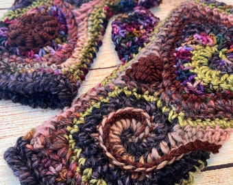 May include: A pair of crocheted fingerless gloves in a multi-coloured yarn with a brown, green, pink, and purple colour scheme. The gloves feature a textured, circular design.