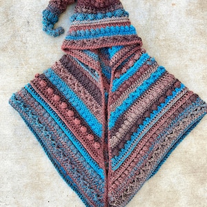 Puede incluir: Una capa con capucha de ganchillo con un sombrero puntiagudo adjunto. La capa está hecha en una variedad de tonos de marrón, azul y turquesa. El sombrero tiene una punta larga y puntiaguda y está decorado con un pompón.