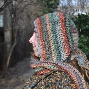 May include: A person wearing a hooded crocheted scarf with a multi-colored striped pattern in shades of brown, green, and orange. The scarf is worn over the person's head and covers their hair.