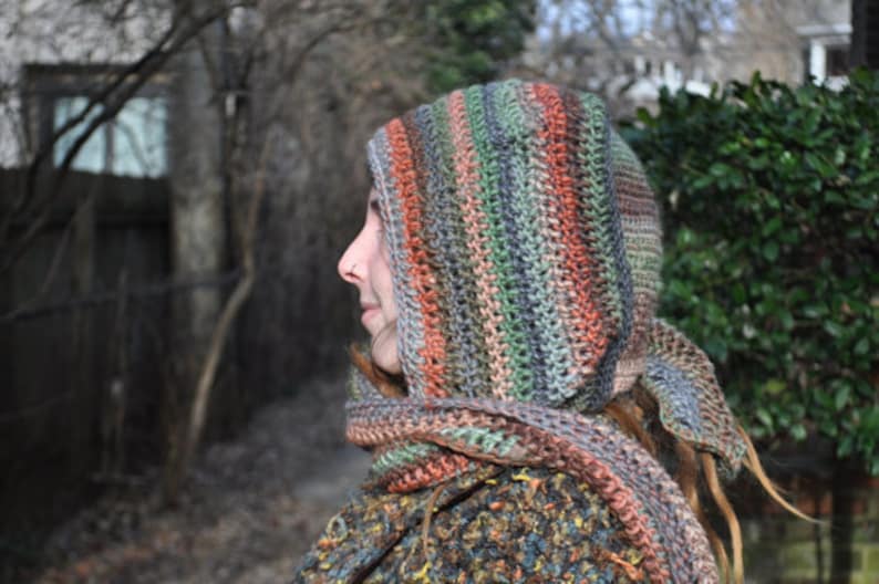 May include: A person wearing a hooded crocheted scarf with a multi-colored striped pattern in shades of brown, green, and orange. The scarf is worn over the person's head and covers their hair.
