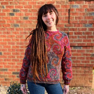 May include: A woman with long brown dreadlocks is wearing a colourful crocheted sweater with a swirling pattern. The sweater is a mix of pink, red, grey, and brown.