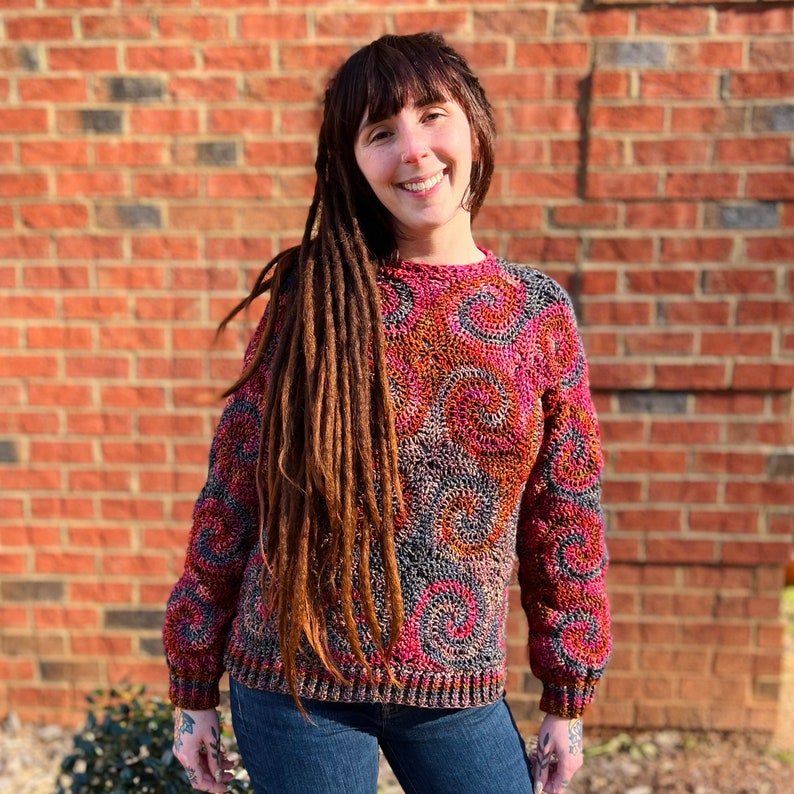 May include: A woman with long brown dreadlocks is wearing a colourful crocheted sweater with a swirling pattern. The sweater is a mix of pink, red, grey, and brown.