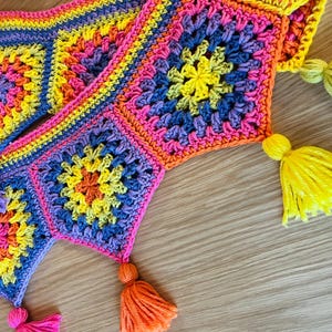 May include: A colorful, handmade crochet scarf featuring a granny square design in vibrant hues of yellow, pink, orange, and blue. The scarf has decorative tassels in coordinating colors at each point. The scarf is displayed on a wooden surface.