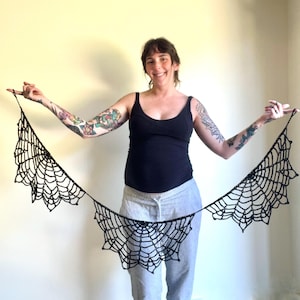 Op de afbeelding: Een zwart gehaakt spinnenwebontwerp, gepresenteerd tegen een lichte achtergrond. Het web is ingewikkeld vervaardigd met een delicate, open weefsel, ideaal voor Halloween of gothic-thema decoratie. Het web wordt horizontaal weergegeven.