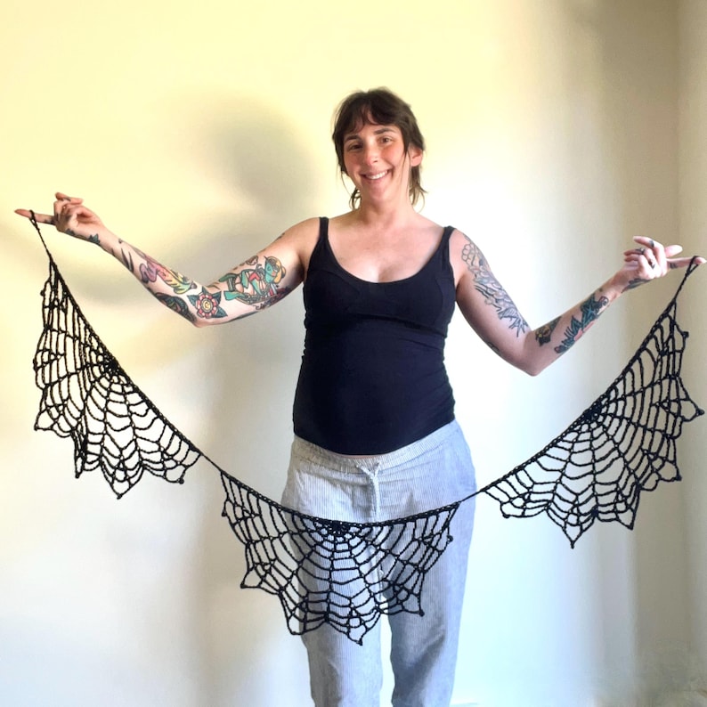Op de afbeelding: Een zwart gehaakt spinnenwebontwerp, gepresenteerd tegen een lichte achtergrond. Het web is ingewikkeld vervaardigd met een delicate, open weefsel, ideaal voor Halloween of gothic-thema decoratie. Het web wordt horizontaal weergegeven.