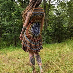 May include: A sleeveless crochet tunic featuring a circular spiderweb pattern in shades of brown, orange, yellow, and blue. The garment has a flared, ruffled hemline and an open-knit construction that displays a dark brown layer underneath.