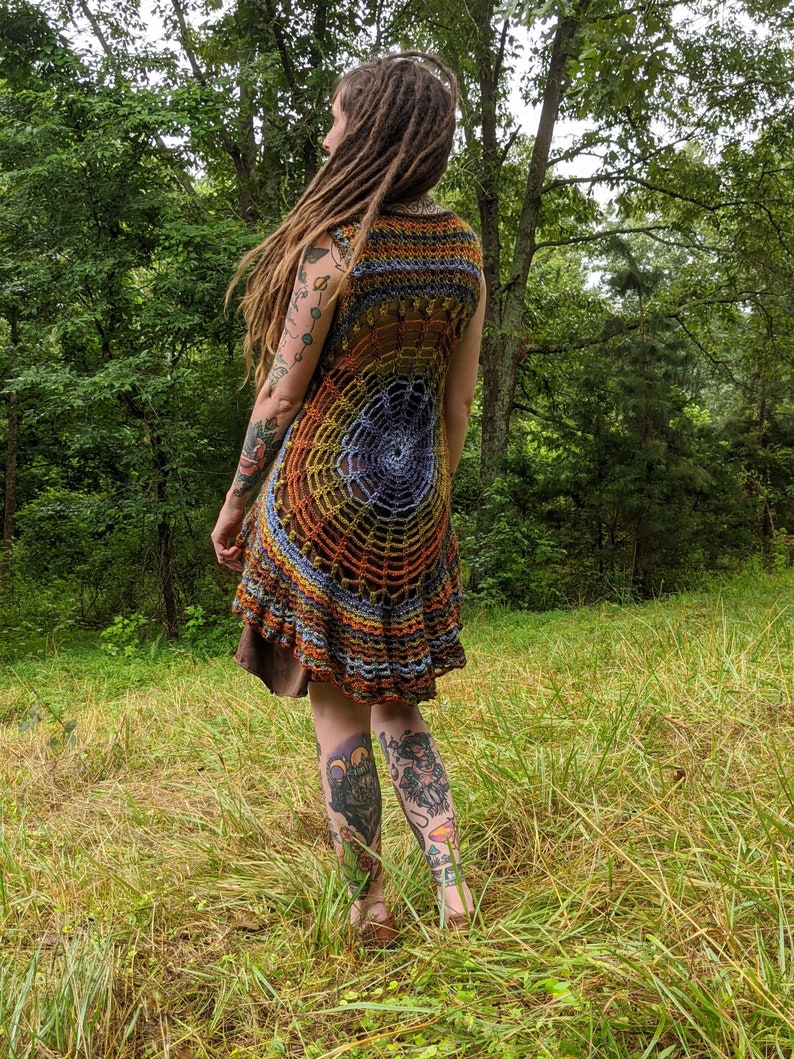 May include: A sleeveless crochet tunic featuring a circular spiderweb pattern in shades of brown, orange, yellow, and blue. The garment has a flared, ruffled hemline and an open-knit construction that displays a dark brown layer underneath.