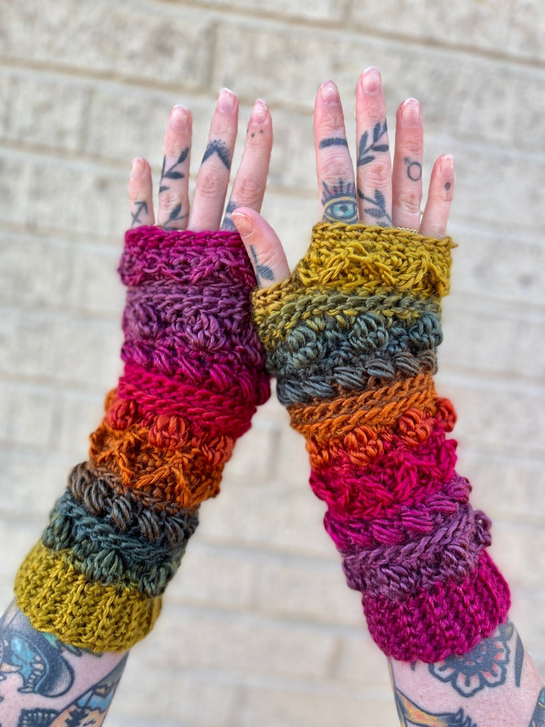 May include: A pair of colorful crocheted fingerless gloves with a textured design. The gloves are made with a gradient of colors, including mustard yellow, orange, red, and purple.