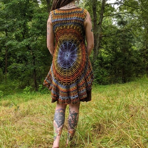 May include: A sleeveless crochet tunic featuring a circular spiderweb pattern in shades of orange, yellow, blue, and purple. The garment has a flared, ruffled hemline and is layered over a solid brown base garment, showcasing intricate yarn work.