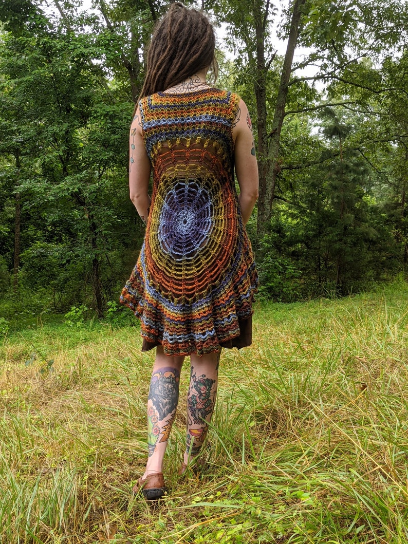 May include: A sleeveless crochet tunic featuring a circular spiderweb pattern in shades of orange, yellow, blue, and purple. The garment has a flared, ruffled hemline and is layered over a solid brown base garment, showcasing intricate yarn work.