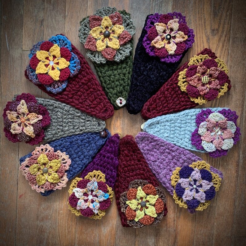 May include: A collection of ten triangular crochet headbands arranged in a circle. Each headband features a unique color combination and a large, multi-layered crocheted flower embellishment at the point, secured with a central button.