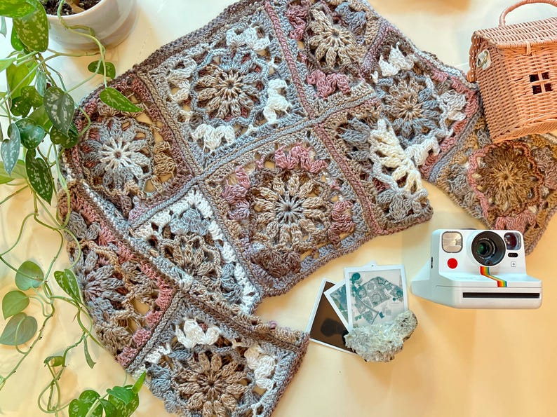 K&ouml;nnte beinhalten: Eine geh&auml;kelte Granny-Square-Decke in Braun-, Rosa- und Wei&szlig;t&ouml;nen. Die Decke besteht aus mehreren Quadraten mit einem Blumenmuster.