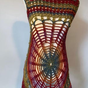 May include: A sleeveless crochet tunic featuring a prominent spiderweb pattern in the center. The garment is crafted from variegated yarn in shades of deep red, olive green, gold, and dark blue, creating a textured, open-work design.
