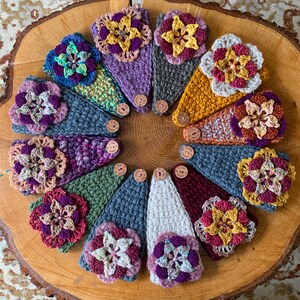 May include: A collection of twelve triangular crochet headbands arranged in a circle. Each headband features a unique color combination, a large crocheted flower embellishment, and a small circular wooden button with a visible logo.