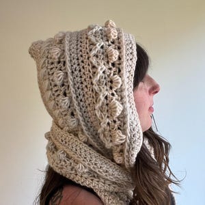Op de afbeelding: Een cr&egrave;mekleurige gehaakte capuchon- en sjaalset. De capuchon heeft een textuurpatroon met verhoogde bobbles en een puntige bovenkant. De sjaal heeft een bijpassende cr&egrave;mekleur en is om de nek gewikkeld. De set is gemaakt van zacht, getextureerd garen.
