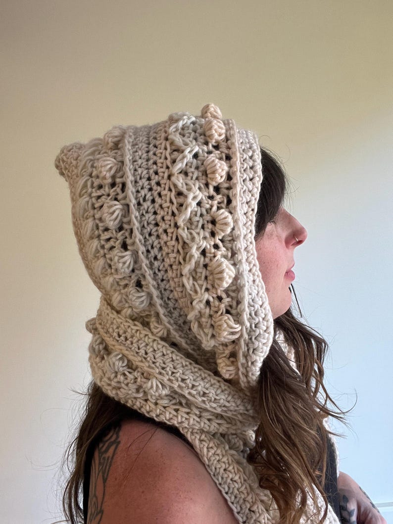Op de afbeelding: Een cr&egrave;mekleurige gehaakte capuchon- en sjaalset. De capuchon heeft een textuurpatroon met verhoogde bobbles en een puntige bovenkant. De sjaal heeft een bijpassende cr&egrave;mekleur en is om de nek gewikkeld. De set is gemaakt van zacht, getextureerd garen.